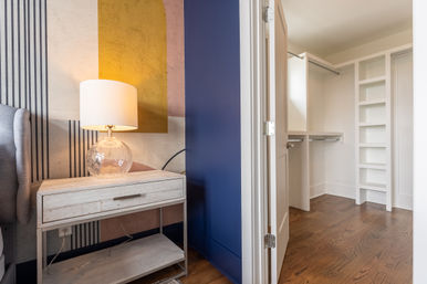Modern bedroom corner with a lit glass table lamp on a distressed white nightstand, geometric accent wall, navy trim, and an open walk-in closet with built-in shelves and hardwood floors.