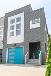 Contemporary gray townhouse exterior with bright turquoise garage and matching front door, white-trimmed windows, small welcoming porch with wreath and 'welcome' sign, driveway in an urban residential setting.