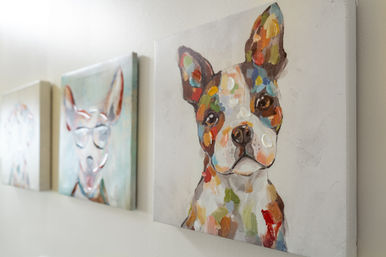 Playful colorful dog portrait on canvas hung on a white interior wall with two blurred companion dog paintings — modern home decor wall art