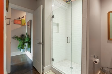 Bright contemporary bathroom with frameless glass walk-in shower tiled in white subway and hexagon floor tiles, open door revealing hallway with potted palm and wall mirror.