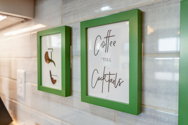 Green-framed kitchen wall decor on a gray tile backsplash: playful sign reading 'Coffee 'till Cocktails' next to a framed coffee cup illustration.