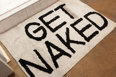 Playful white shag bath mat with bold black letters reading GET NAKED, set on a brown bathroom floor beside a white tiled tub.