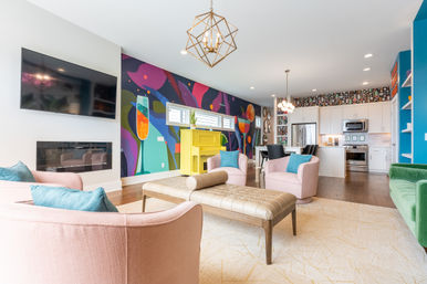 Bright open-concept living room and kitchen with a colorful abstract mural, yellow piano, pink armchairs, tufted leather bench, green sofa, wall-mounted TV and white kitchen with stainless appliances