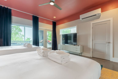 Modern bedroom with two white-made beds topped with folded towels, coral-red ceiling with fan, wall-mounted TV, air conditioner, and sliding glass doors opening to a tree-lined balcony.