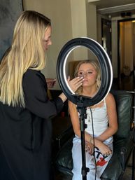Makeup artist applying finishing touches to a seated blonde woman under a ring light during an indoor beauty session