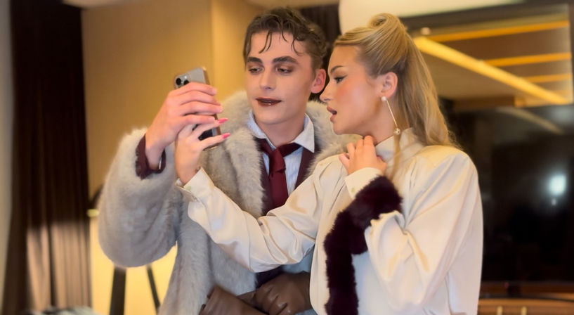 Two stylish young adults in fur-trimmed coats looking at a smartphone together in a modern luxury hotel suite — glamorous evening fashion moment.
