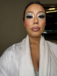 Close-up indoor portrait of a person in a white plush robe showcasing glam makeup: shimmery blue and silver eyeshadow, dramatic false lashes, rosy cheeks, and glossy mauve lips under vanity-style lighting.