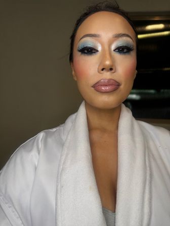 Close-up indoor portrait of a person in a white plush robe showcasing glam makeup: shimmery blue and silver eyeshadow, dramatic false lashes, rosy cheeks, and glossy mauve lips under vanity-style lighting.