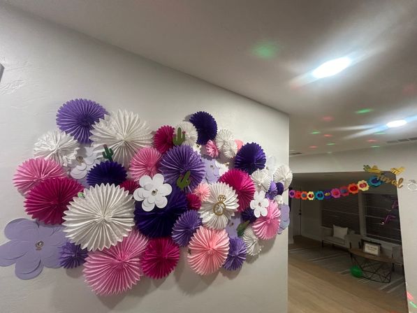 Colorful party backdrop of pink, purple and white paper fan rosettes and paper flowers on a home hallway wall, with a floral garland and living room seating visible in the background.