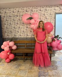 Smiling pregnant woman in a flowy hot-pink dress posing by a pink balloon garland and wooden bench against a floral wallpaper backdrop at an indoor baby shower