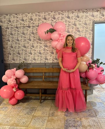 Smiling pregnant woman in a flowy hot-pink dress posing by a pink balloon garland and wooden bench against a floral wallpaper backdrop at an indoor baby shower