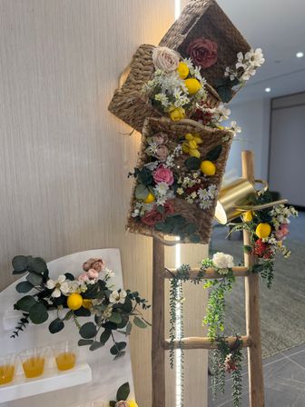 Rustic wooden ladder display with woven wicker baskets of faux roses, daisies, eucalyptus and bright decorative lemons, brass watering can and trailing greenery against a light wall, small clear cups of orange drink on a nearby shelf — indoor spring floral decor.