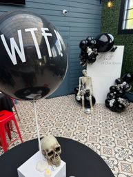 Spooky-chic indoor event setup with a large black "WTF!" balloon over a skull table prop, a seated skeleton beside a white photo backdrop, black balloon garlands and patterned tiled floor.