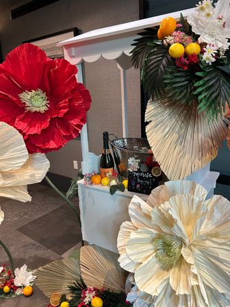 White pop-up drink cart with a champagne bottle in an ice bucket, surrounded by oversized red and cream paper flowers, palm fronds, dried fan palms and bright citrus accents — colorful event decor