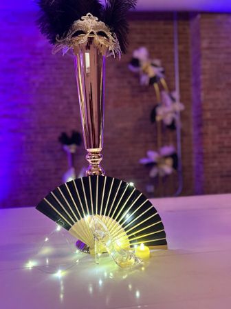 Masquerade-themed table centerpiece with a tall gold vase topped by a feathered mask, black-and-gold folding fan, clear glass slipper, warm fairy lights and candle against purple uplighting and brick backdrop