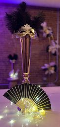 Tall gold vase centerpiece topped with an ornate silver masquerade mask and black feathers, paired with a black-and-gold folding fan, fairy lights and a glass slipper — elegant masquerade table decor.