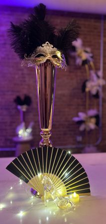 Tall gold vase centerpiece topped with an ornate silver masquerade mask and black feathers, paired with a black-and-gold folding fan, fairy lights and a glass slipper — elegant masquerade table decor.