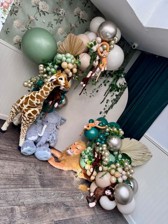 Playful safari-themed kids party backdrop with green, cream and metallic balloons, faux greenery and dried palm accents, and plush giraffe, elephant, lion and monkeys.