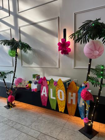 Indoor Hawaiian-themed party display with colorful 'ALOHA' surfboard signs, faux palm trees, pink paper lanterns and flamingo decorations
