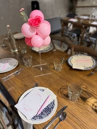 Playful pink balloon cluster with a faux rose on a clear stand, centered on a wooden dining table set with blue-and-white plates, pink pens, glass tumblers, and silverware — rustic indoor celebration table setting.