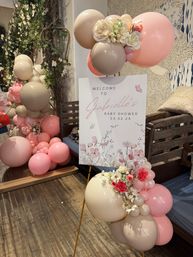 Pastel floral baby shower welcome sign on easel, surrounded by pink and beige balloon garlands and faux roses in a cozy indoor venue