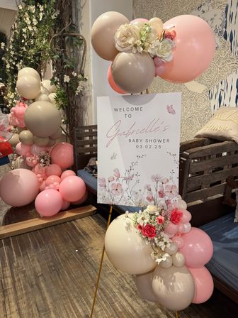 Pastel floral baby shower welcome sign on easel, surrounded by pink and beige balloon garlands and faux roses in a cozy indoor venue