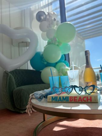 Sunlit bridal celebration table with teal-and-mint balloon garland, bottle of sparkling wine, blue "I do crew" cups and a "Bride" cup, heart-shaped sunglasses on a Miami Beach book.