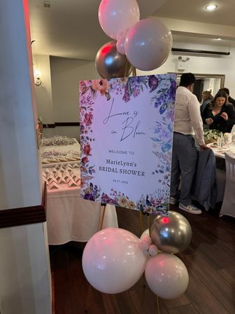 Indoor bridal shower welcome sign with floral "Love is in Bloom" design on an easel, pink and metallic balloons clustered at the base, dessert table and guests in the background at a reception.