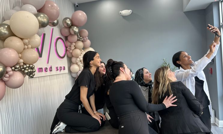 Smiling med spa staff taking a group selfie in a bright reception area decorated with a pink and metallic gold balloon arch and soft gray walls.