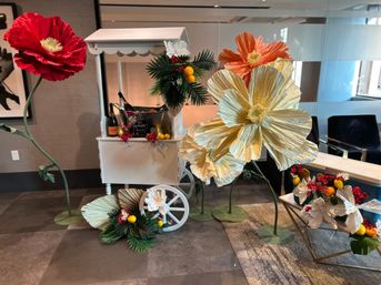 Decorative white cart in a modern lobby featuring oversized paper flowers in red, orange and cream, faux palm leaves, bright lemons and a champagne bucket — colorful floral display
