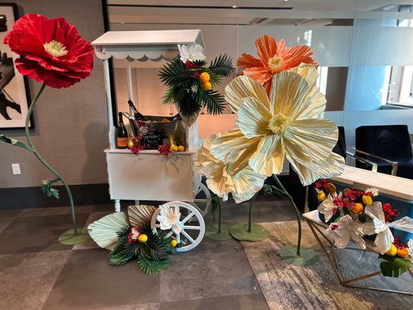 Decorative white cart in a modern lobby featuring oversized paper flowers in red, orange and cream, faux palm leaves, bright lemons and a champagne bucket — colorful floral display