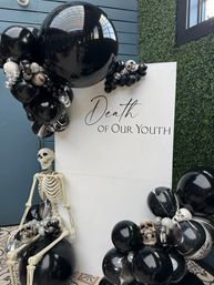Indoor Halloween-themed backdrop with white sign reading 'Death of Our Youth', black and silver balloon garlands, skulls, and seated skeleton.