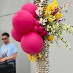 Playful outdoor decoration: cluster of bright magenta balloons and yellow spring flowers tied to a rope-wrapped post, with a blurred person in sunglasses in the background.
