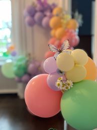 Close-up of a pastel balloon garland in pink, green, yellow and lavender, accented with small white daisies and a delicate butterfly for indoor party decor.