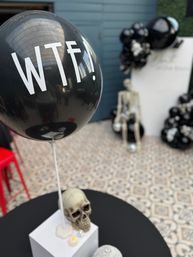 Quirky Halloween-style event decor: black balloon reading 'WTF!' floating above a skull centerpiece, with a seated skeleton and black-and-white balloon clusters in an indoor venue on patterned tile floor.