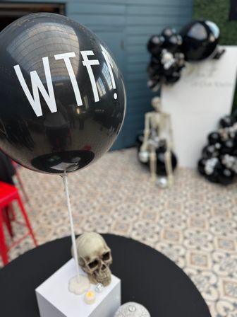 Quirky Halloween-style event decor: black balloon reading 'WTF!' floating above a skull centerpiece, with a seated skeleton and black-and-white balloon clusters in an indoor venue on patterned tile floor.