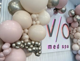 Cheerful blush, cream and metallic balloon garland framing a sleek white med spa wall sign with soft pink LED glow and textured wall panels.
