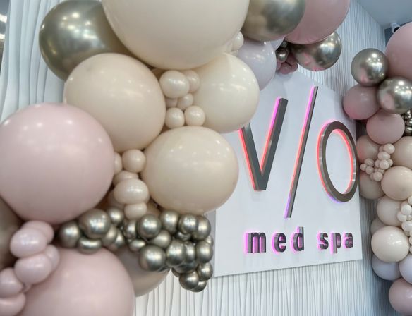 Cheerful blush, cream and metallic balloon garland framing a sleek white med spa wall sign with soft pink LED glow and textured wall panels.