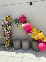 Outdoor patio decoration: a playful diagonal garland of bright pink, orange and yellow balloons woven with white and pastel flowers and faux lemons above three round concrete stools against a beige wall.