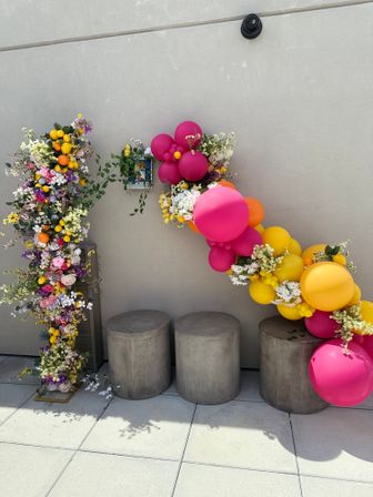 Outdoor patio decoration: a playful diagonal garland of bright pink, orange and yellow balloons woven with white and pastel flowers and faux lemons above three round concrete stools against a beige wall.