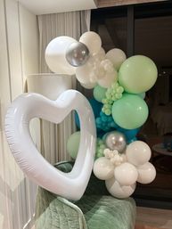 Pastel balloon garland in mint, teal, white and silver wrapped around a large white heart-shaped inflatable, staged indoors on a green velvet chair — modern party decor.