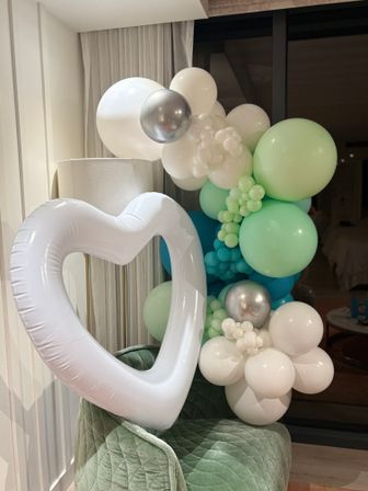Pastel balloon garland in mint, teal, white and silver wrapped around a large white heart-shaped inflatable, staged indoors on a green velvet chair — modern party decor.