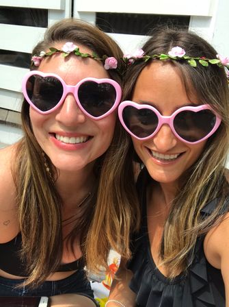 Close-up selfie of two smiling friends wearing pink heart-shaped sunglasses and floral crowns at a sunny outdoor summer gathering