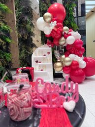 Valentine's candy bar display in an indoor event space: red, white and gold balloon garland with heart accents, jars of striped candy and a tiered shelf of champagne flutes against a vertical green wall.