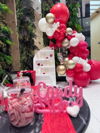 Valentine's candy bar display in an indoor event space: red, white and gold balloon garland with heart accents, jars of striped candy and a tiered shelf of champagne flutes against a vertical green wall.