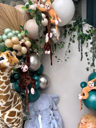 Jungle safari kids' party backdrop with plush giraffe and elephant, hanging monkey toys, teal, cream and metallic balloon garland, dried palm fan and trailing faux greenery