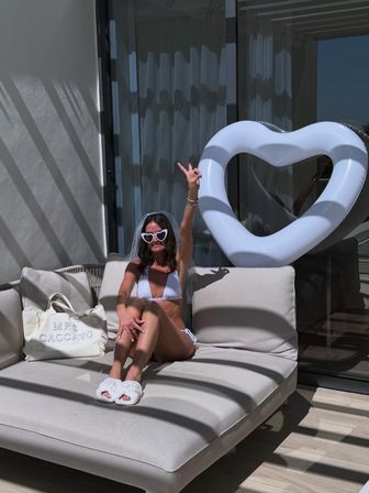 Sunny poolside resort patio — woman in a white bikini with heart-shaped sunglasses and veil lounges on a beige daybed, flashing a peace sign next to an oversized white heart float.
