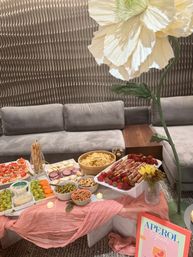Whimsical indoor cocktail party setup: charcuterie spread on a low marble table with grapes, carrots, olives, crackers, chips, almonds and prosciutto-wrapped strawberries, pink draped linen, oversized paper flower decor, Aperol Spritz sign and grey lounge seating with textured wall backdrop.