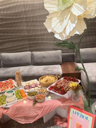 Whimsical indoor cocktail party setup: charcuterie spread on a low marble table with grapes, carrots, olives, crackers, chips, almonds and prosciutto-wrapped strawberries, pink draped linen, oversized paper flower decor, Aperol Spritz sign and grey lounge seating with textured wall backdrop.
