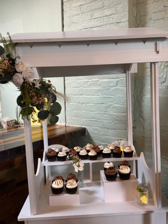 White wooden two-tier cupcake stand against a painted brick wall, featuring assorted chocolate and vanilla cupcakes with drizzled frosting, accented by white roses, eucalyptus greenery, small glass vases and a lit votive for an indoor dessert display